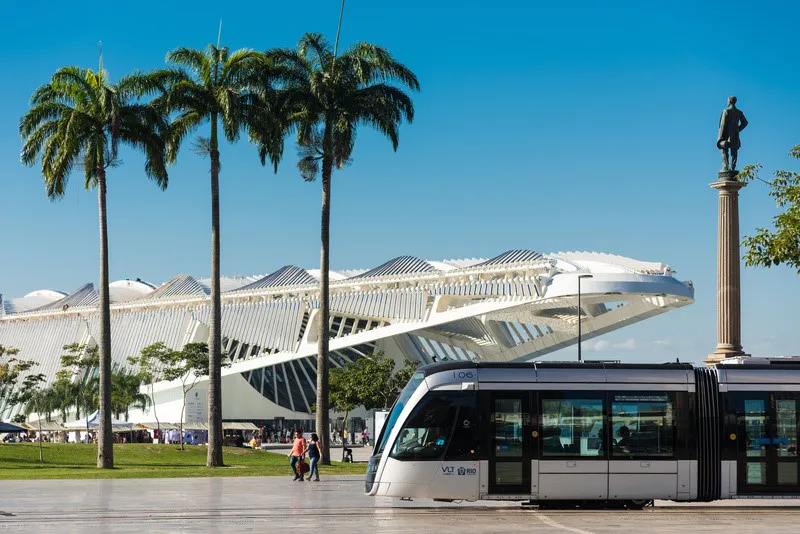  VLT circulando na região portuária do Rio de Janeiro, em frente ao Museu do Amanhã, com palmeiras e monumento histórico ao fundo