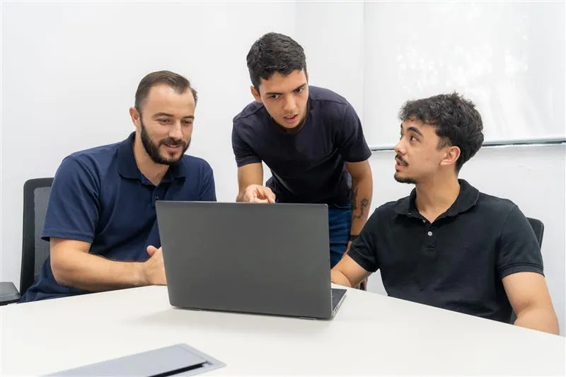  Três homens analisando a tela de um laptop, envolvidos em uma conversa.