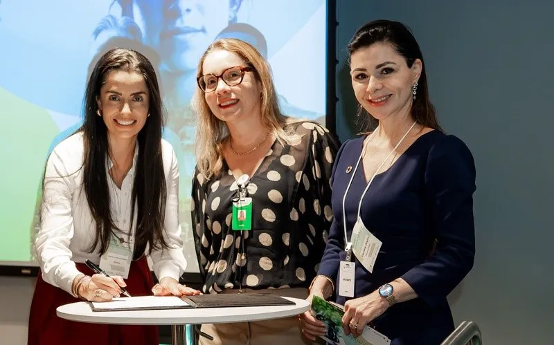 Três mulheres sorrindo diante de uma mesa com documentos, em ambiente com telão de apresentação ao fundo.