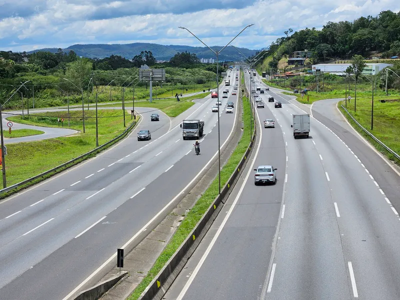 Rodovia movimentada com carros e caminhões em ambos os sentidos, cercada por área verde e céu nublado.
