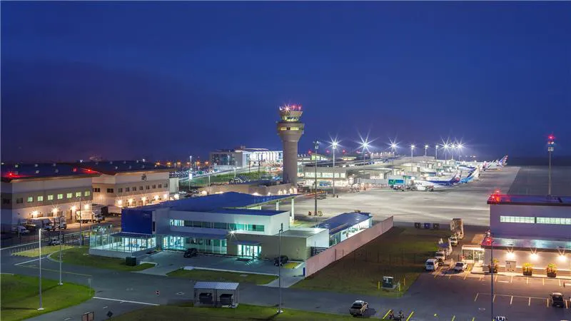  Vista noturna de um aeroporto iluminado, com torre de controle, terminal de embarque e vários aviões alinhados no pátio.