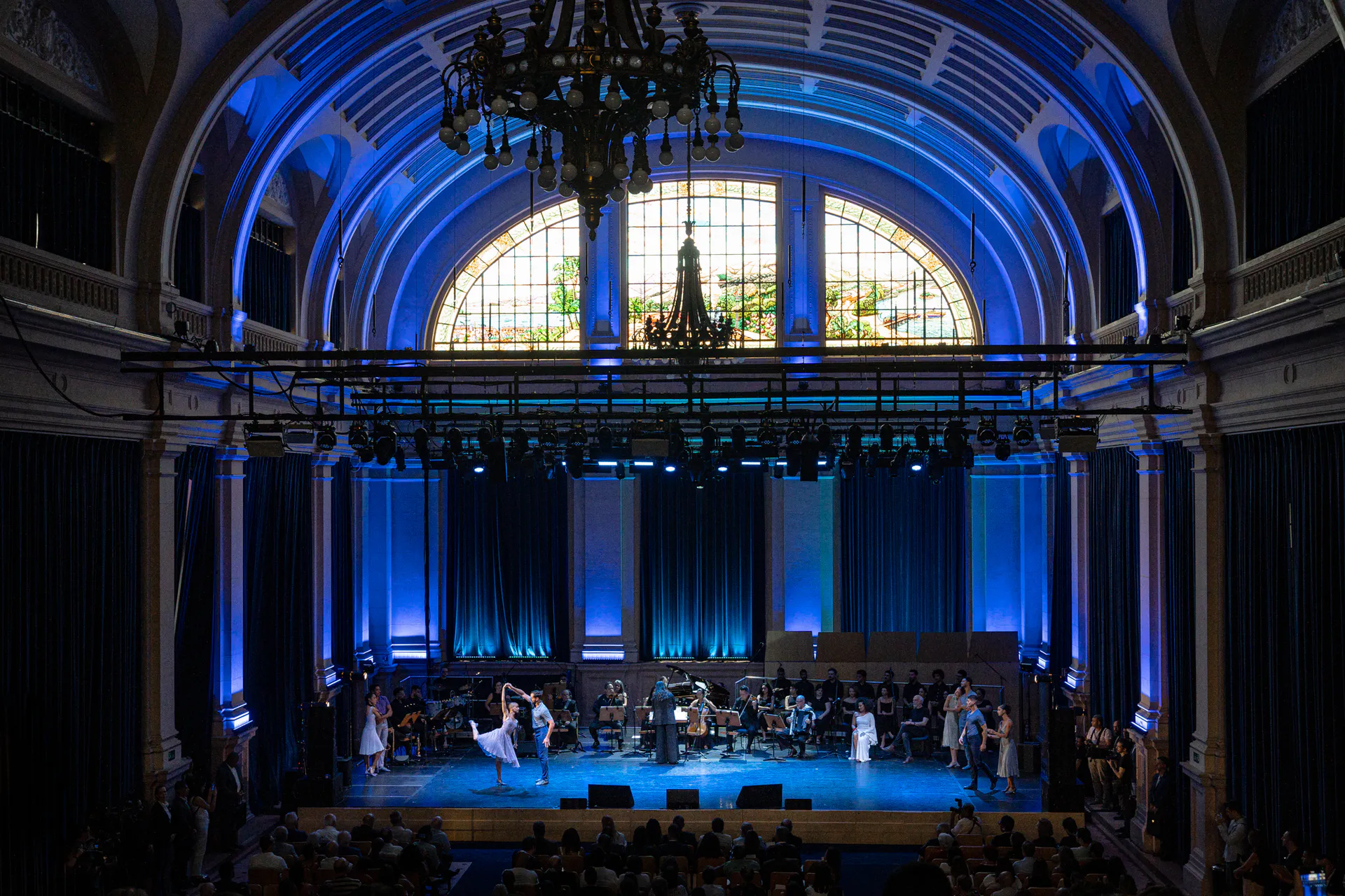 Espetáculo com orquestra e bailarinos no palco da Sala São Paulo, com plateia e iluminação azul no ambiente.