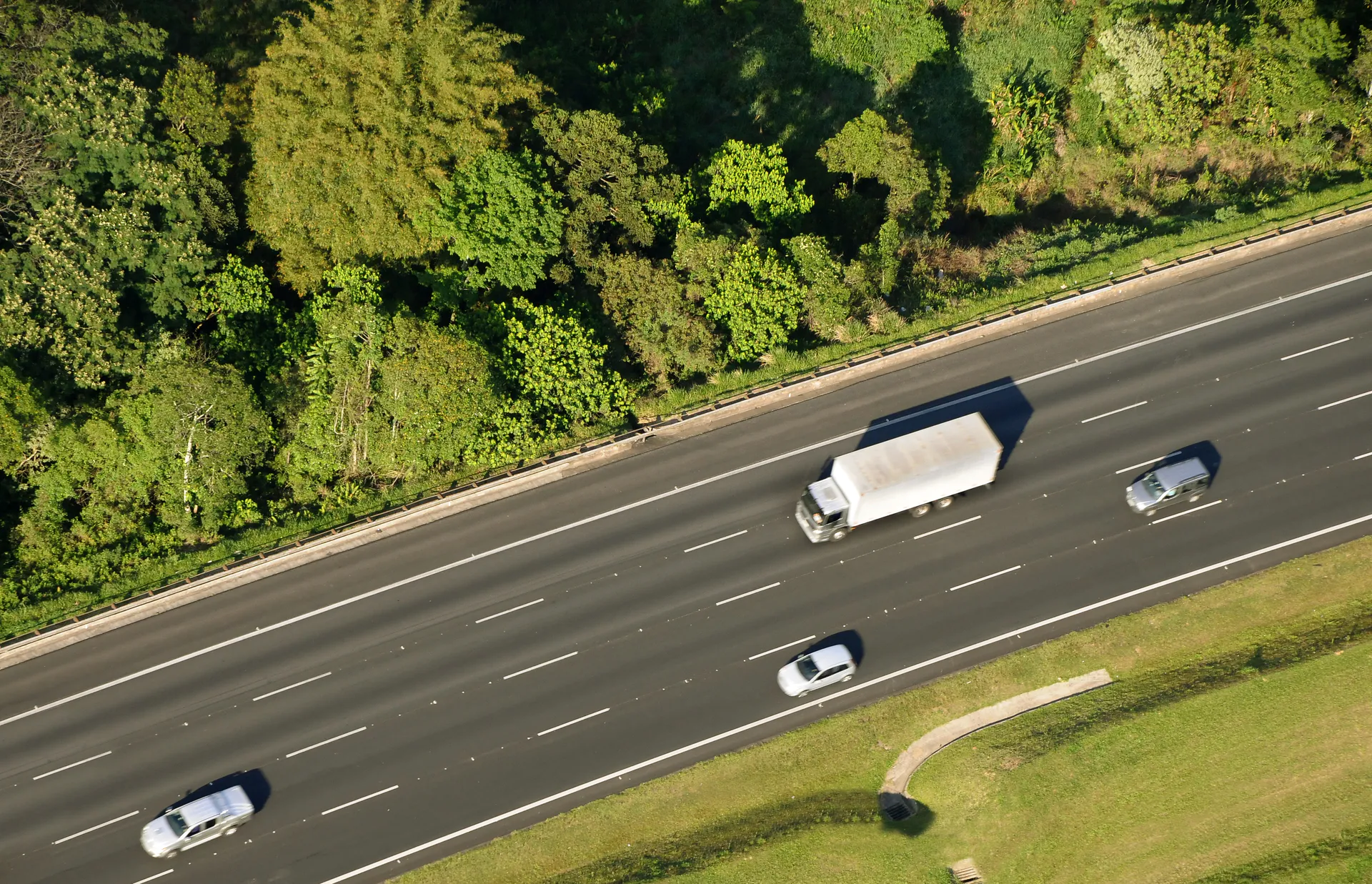 Vista aérea de um caminhão e dois carros andando por uma rodovia ladeada por vegetação
