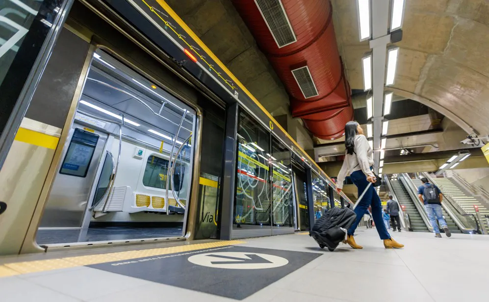  Passageira com mala de rodinhas aguarda embarque em plataforma de metrô moderna, com trem parado e portas automáticas abertas.