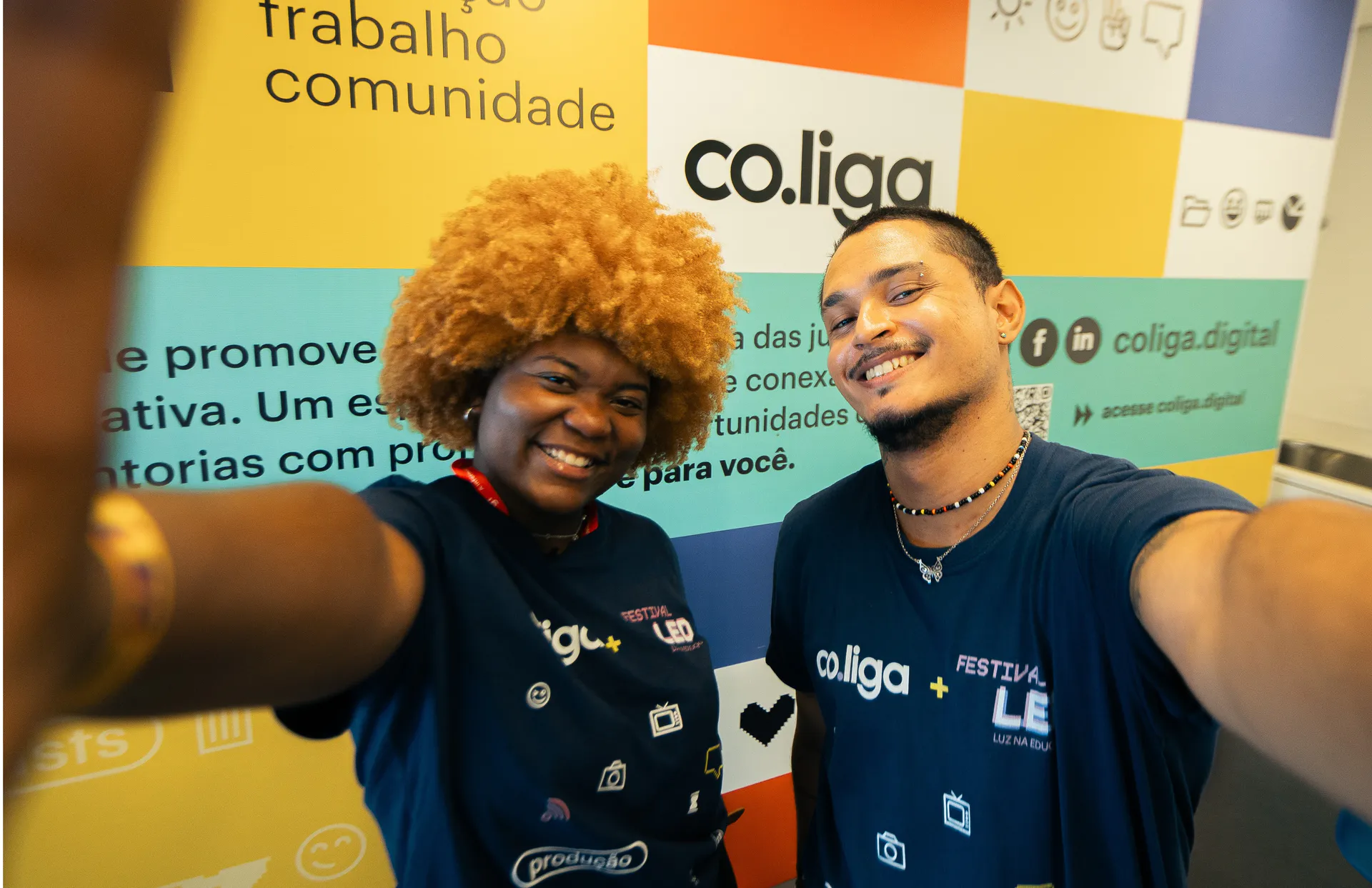  Jovens participantes da co.liga sorrindo em selfie durante evento, com painel colorido da plataforma ao fundo.