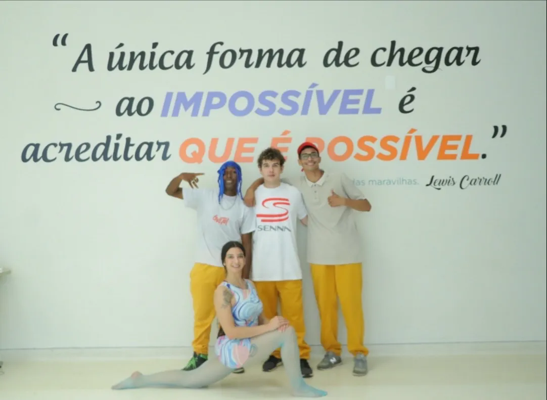 Grupo de jovens artistas em frente a parede com frase inspiradora; uma bailarina está à frente em posição de alongamento.